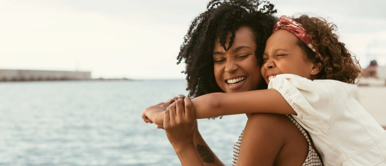 Smiling mother holding small child on her back with view of water in the background